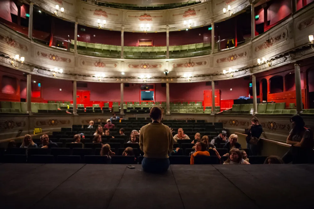 A person sits on the edge of a stage and speaks to an audience in an old-fashioned theatre auditorium.
