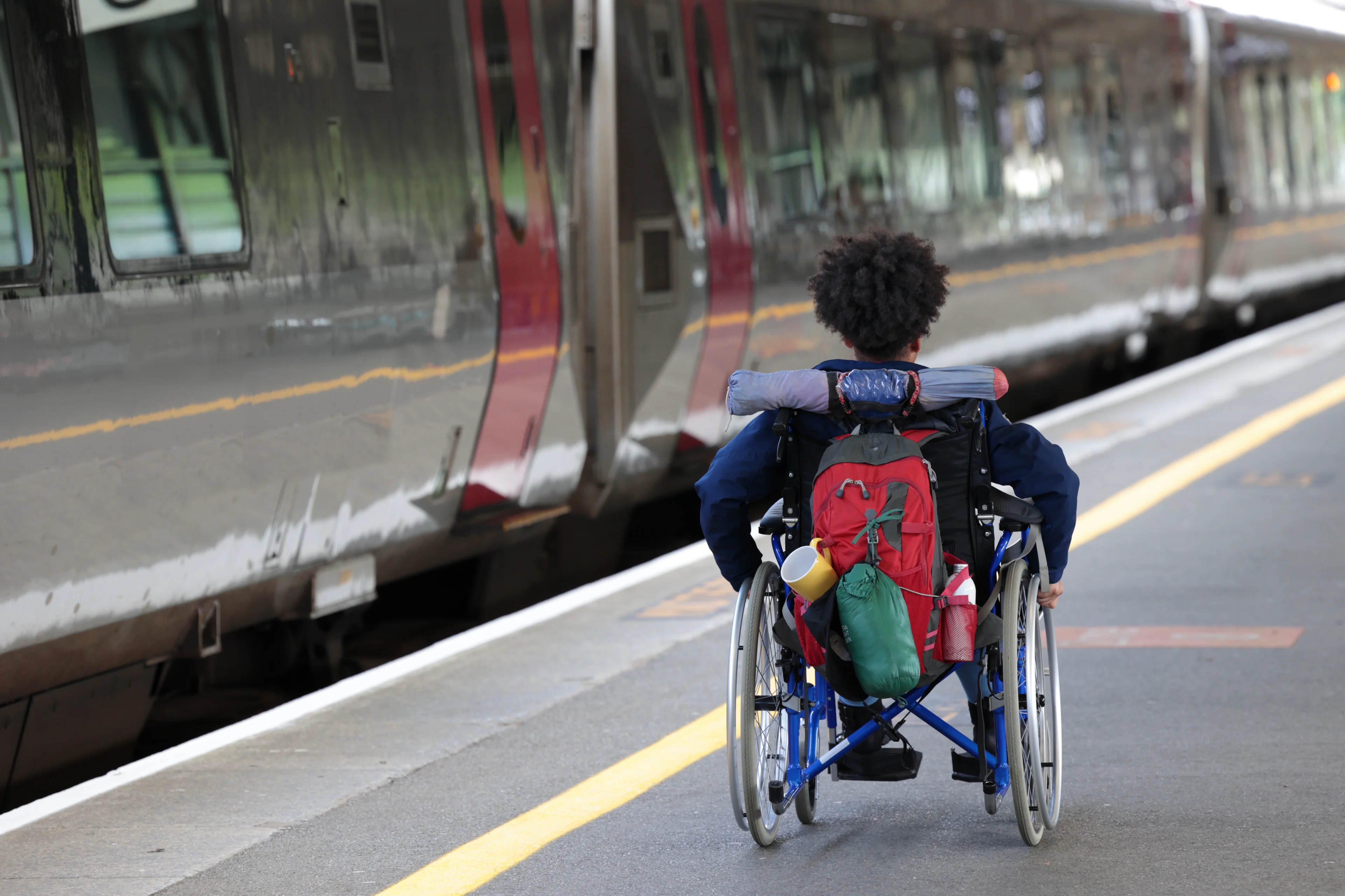 A woman in a wheelchair with camping equipment on the back wheels herself down a train platform with a train standing next to it.