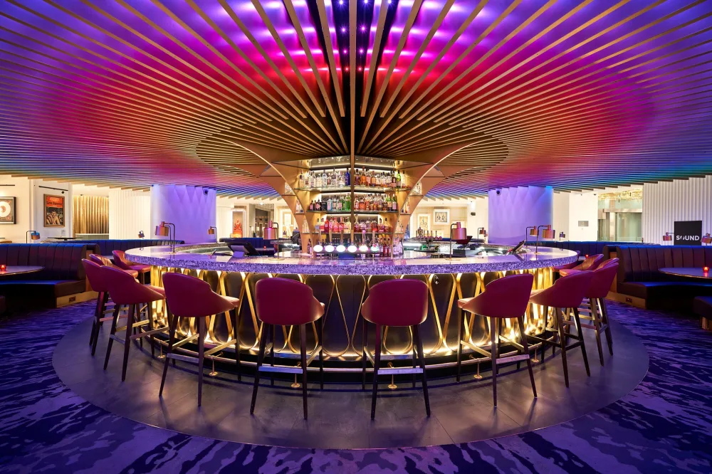 A funky circular gold hotel bar with red and purple lights above it and red high back chairs around it. 