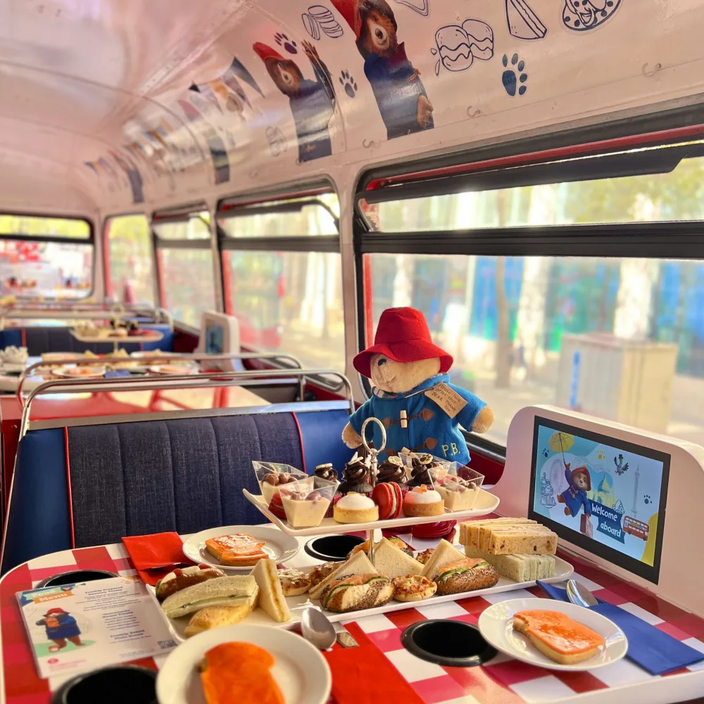 Afternoon tea spread onto a table on the top deck of a double decker bus, with a cuddly toy Paddington bear.