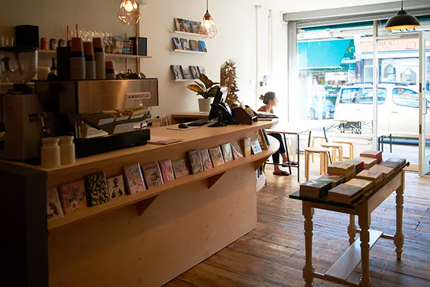 A stylish bookshop cafe with wooden floors and a white woman at a table looking out the window.