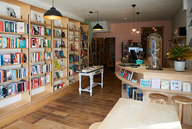 The interior of a stylish bookshop cafe with a wooden floors, floor to ceiling shelves on one side and a bar/counter on the other.