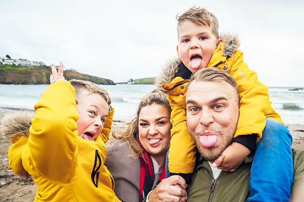 A white man and a white woman, along with 2 young white boys in matching yellow anoraks, standing on a beach on a wintry day and sticking their tongues out.